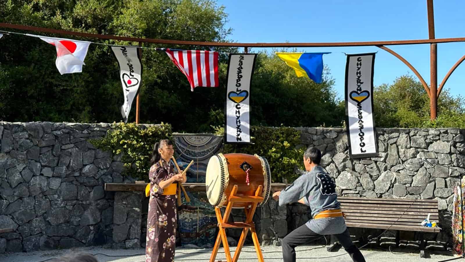 Two people in traditional Japanese dress play a drum in front of Japanese, American and Ukrainian flags.