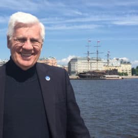 Tim Quigley smiling in a dark jacket standing by a waterfront with a bridge and a ship in the background.