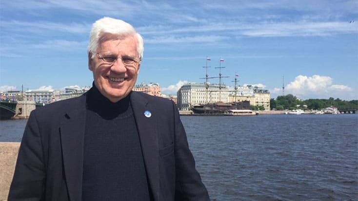 Tim Quigley smiling in a dark jacket standing by a waterfront with a bridge and a ship in the background.