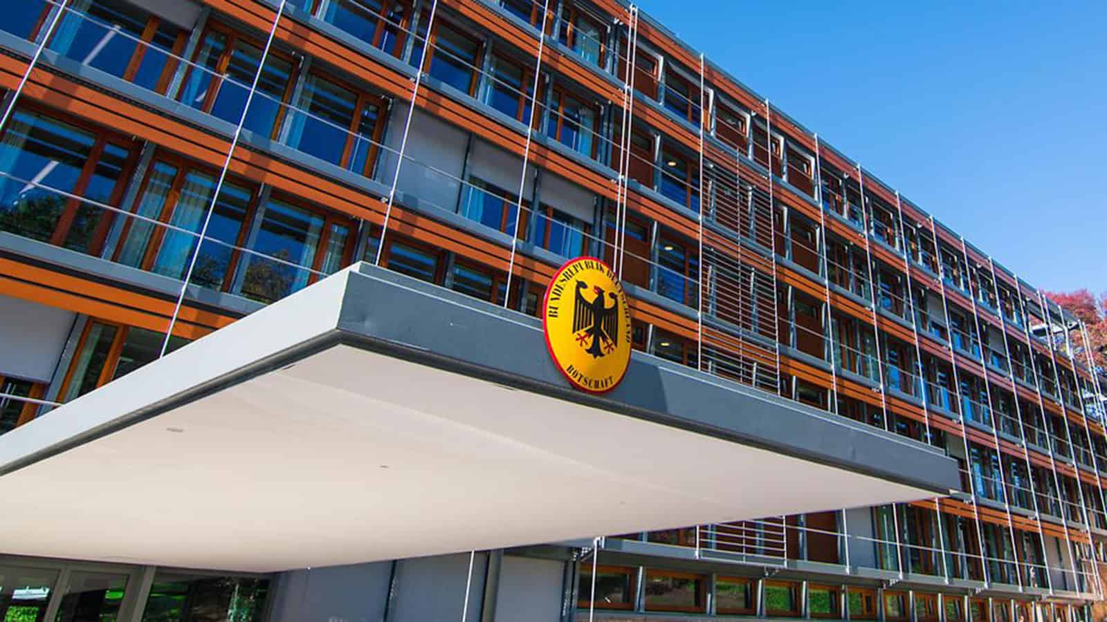 The German Embassy in Washington DC has a modern building exterior, with its glass windows and prominent emblem of a black eagle on a yellow background above the entrance.