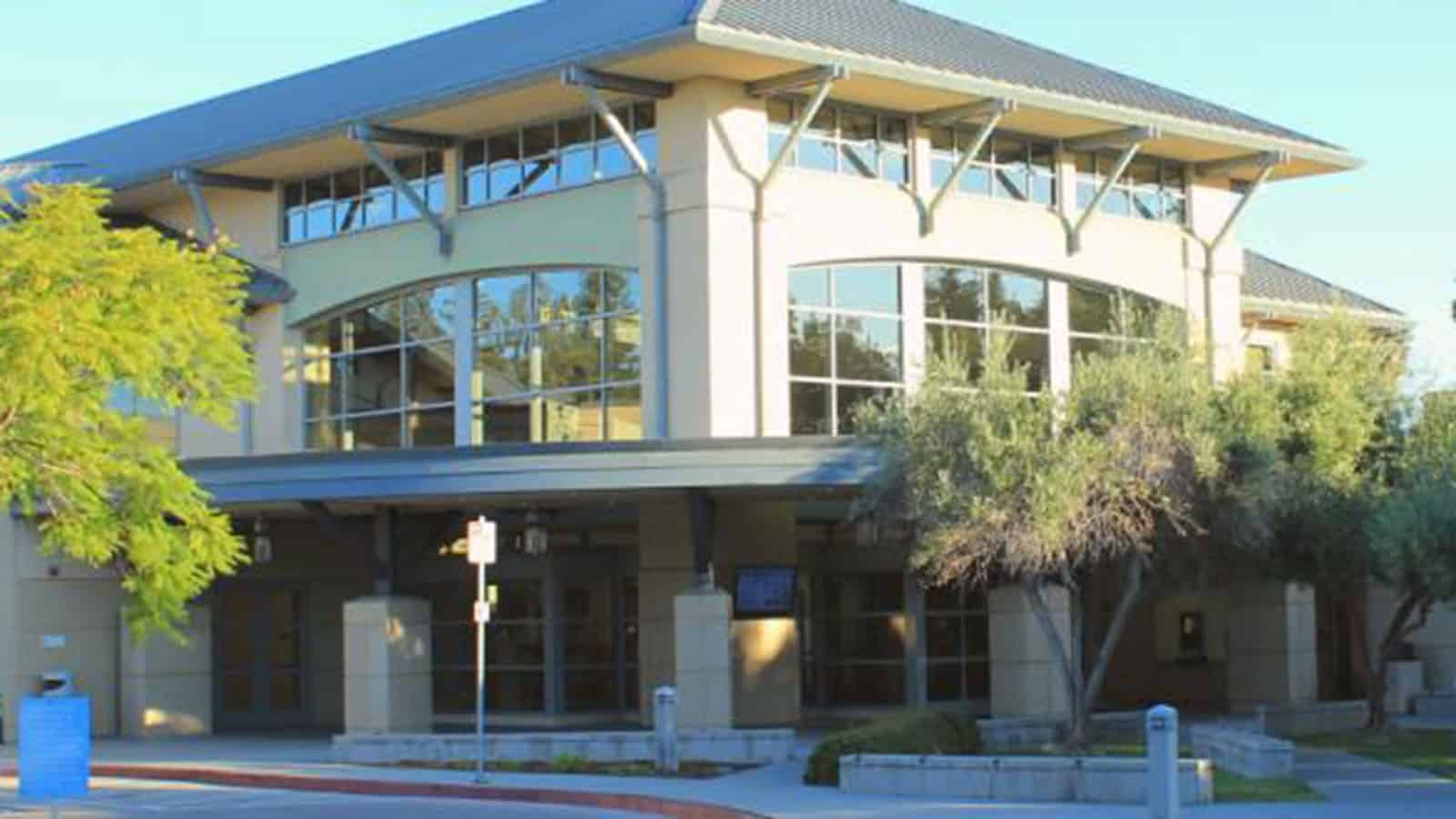 The Santa Clara Central Park Library is a modern, two-story building with large windows and a sloped roof, surrounded by trees and a paved walkway.