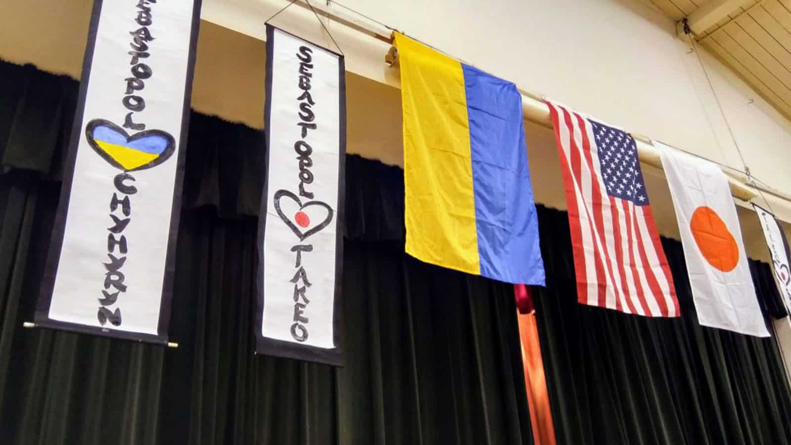 Flags of Ukraine, United States, and Japan hang alongside banners with the names of Chyhyryn and Takeo, featuring heart designs, in an auditorium setting for a Friendship Celebration. The ambiance is enriched with the aroma of borshch and sushi.