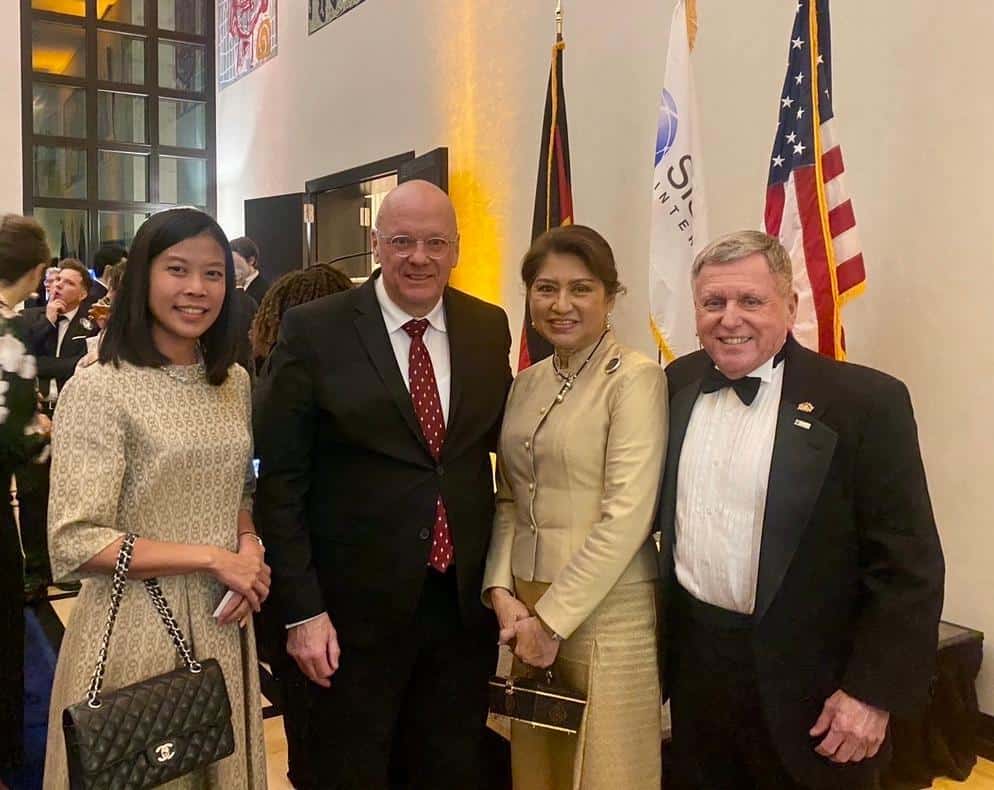 Four people in formal attire stand together at an indoor event. Two women are in dresses, and two men wear suits. U.S. flags are visible in the background. They are all smiling.