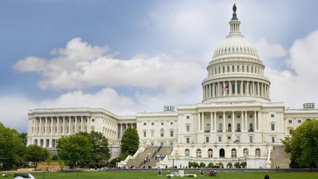 The United States Capitol building with its dome and adjacent wings, set against a partly cloudy sky, surrounded by trees and a lawn with people.