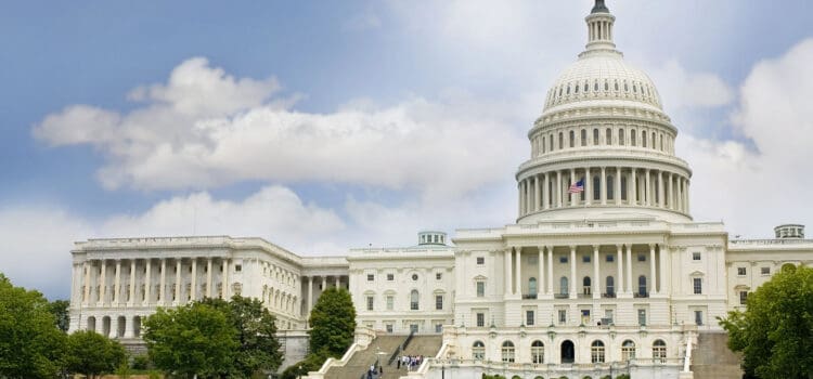 The United States Capitol building with its dome and adjacent wings, set against a partly cloudy sky, surrounded by trees and a lawn with people.