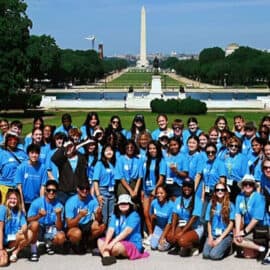 A large group of people in matching blue shirts pose for a photo outside at the 2025 Youth Leadership Summit, with the Washington Monument and National Mall visible in the background.