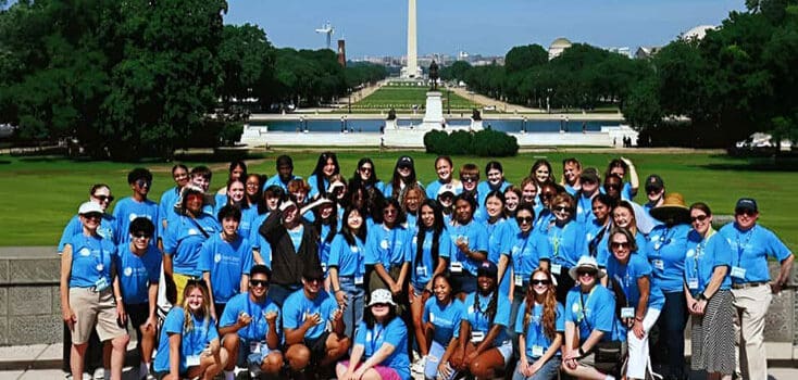 A large group of people in matching blue shirts pose for a photo outside at the 2025 Youth Leadership Summit, with the Washington Monument and National Mall visible in the background.