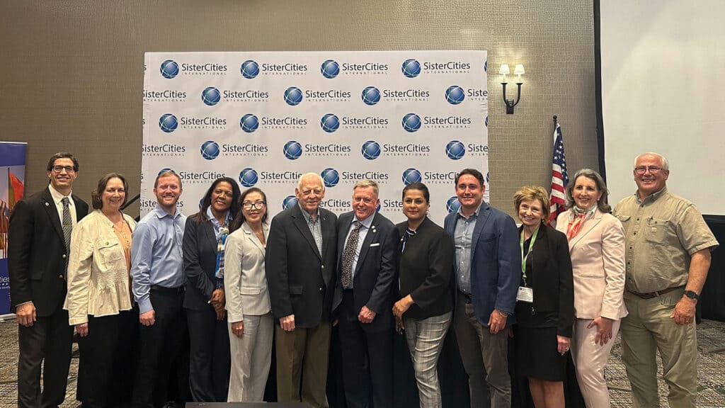 A group of thirteen people poses together in front of a Sister Cities International backdrop at an indoor event.