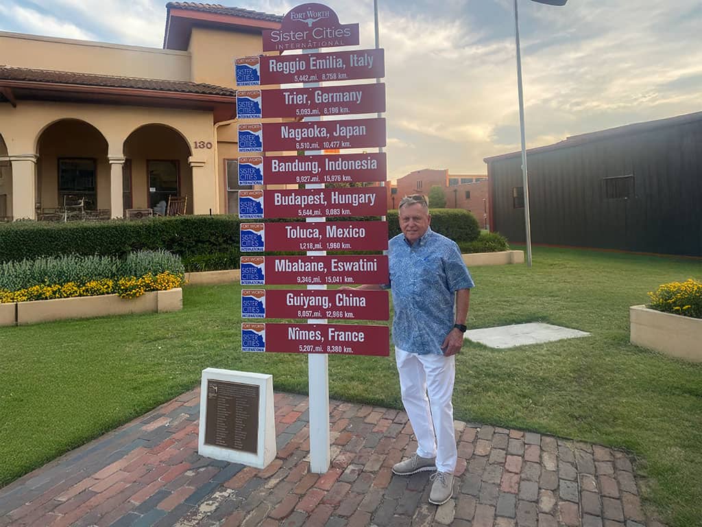 A man stands beside a signpost listing various sister cities and their distances in front of a building and garden area.