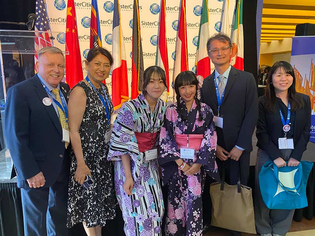 Six people stand together indoors, two wearing traditional Japanese outfits, in front of international flags and a "Sister Cities" banner at an event.
