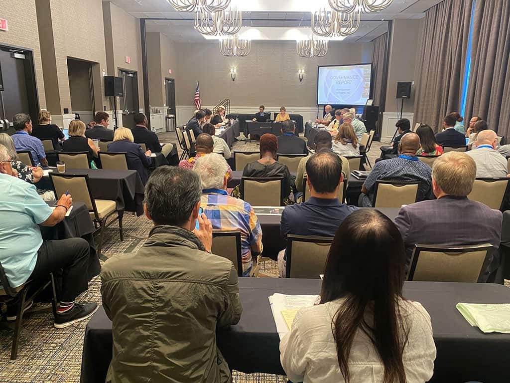 A group of people seated in a conference room listens to a presentation, with a screen at the front displaying a slide.