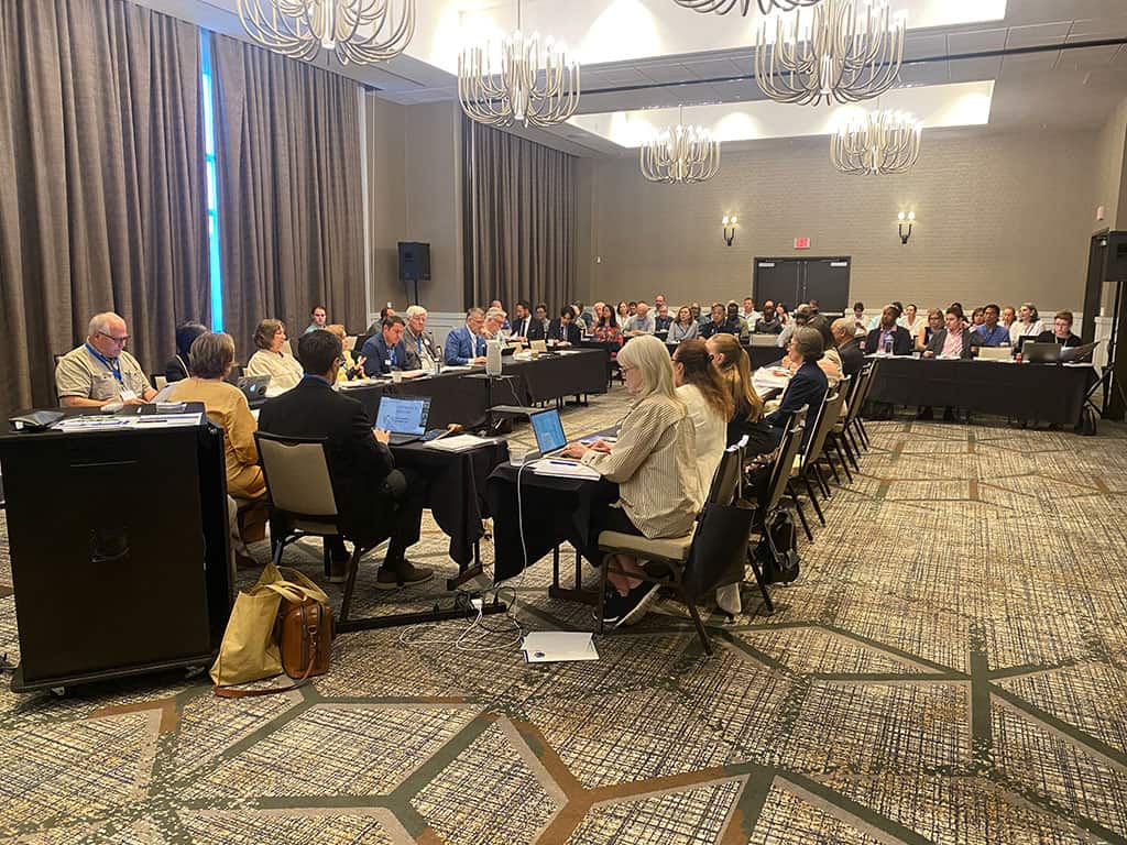 A large group of people sit at tables in a conference room, some working on laptops, while listening to a discussion or presentation.