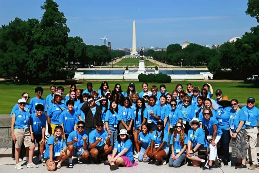 A large group of people in blue shirts pose for a photo in front of the National Mall in Washington, D.C., with the Washington Monument in the background.
