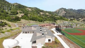 Aerial view of a sports complex with a building, baseball field, and artificial turf, surrounded by hills and greenery under a clear sky.