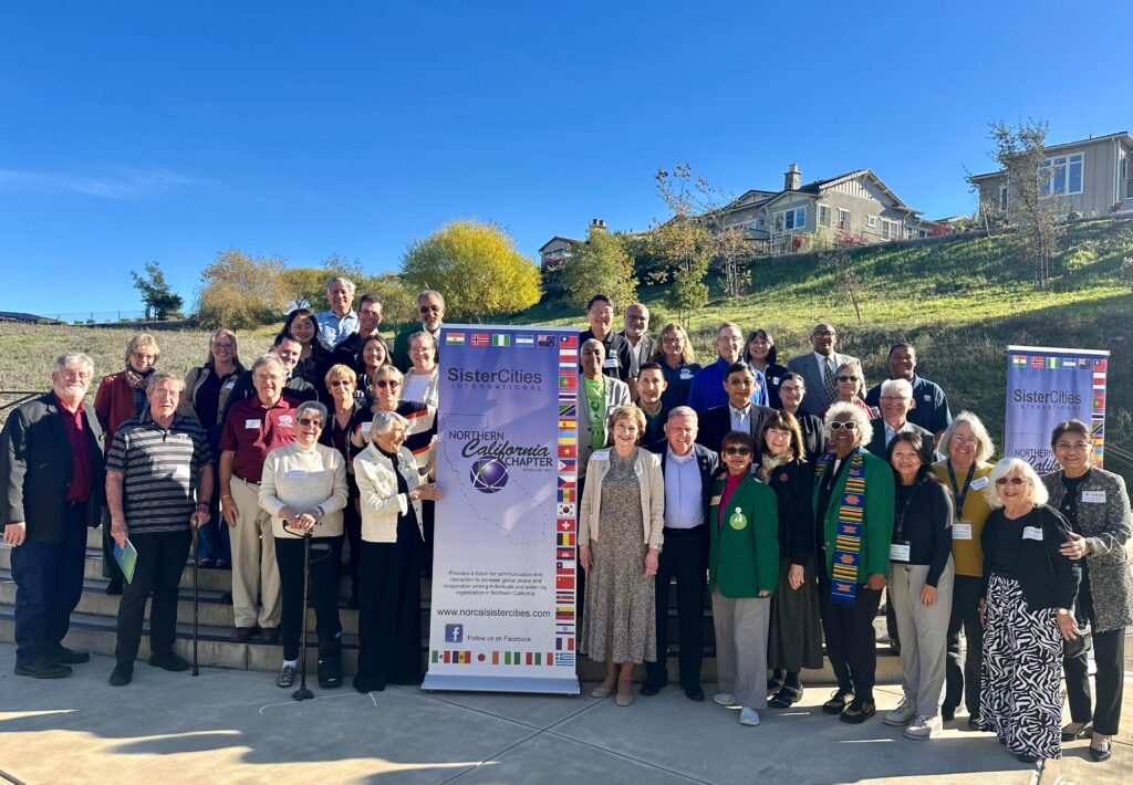 2025 NorCal Sister Cities conference attendees pose outdoors in front of two Sister Cities banners, with houses and a grassy hill in the background on a sunny day.