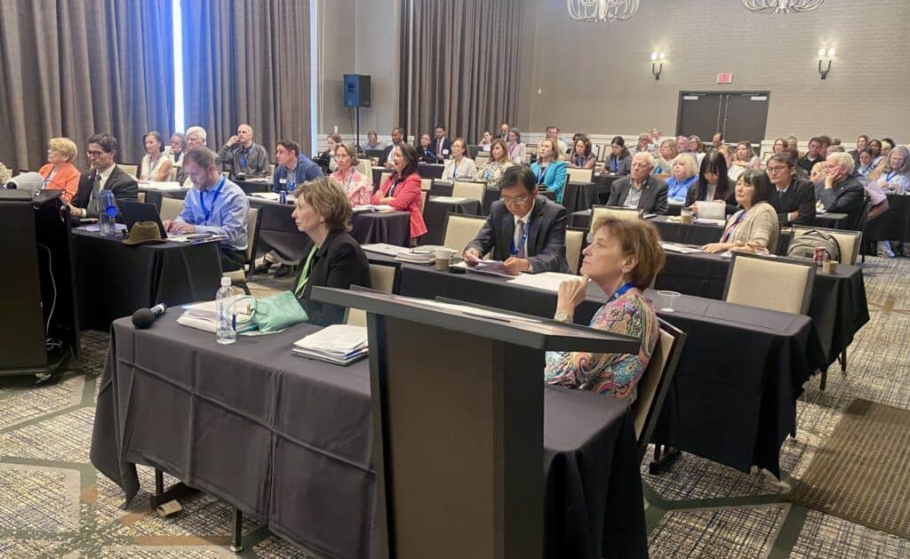 A group of people sit at tables in a conference room, listening to a presentation. Some are taking notes while others look toward the front of the room.