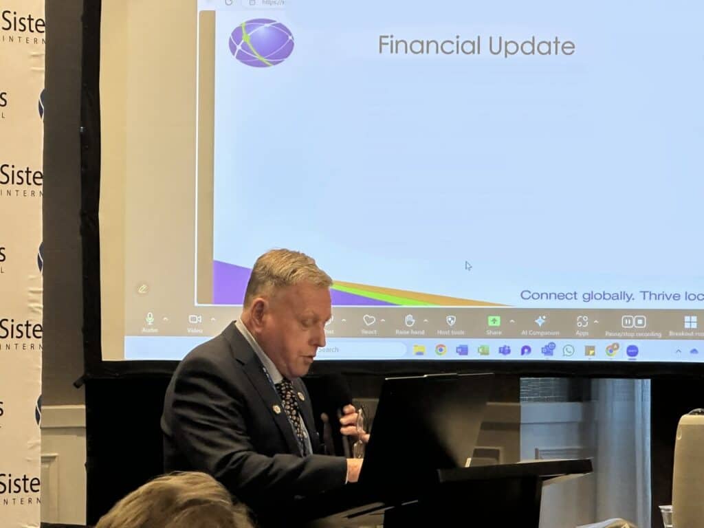 A man in a suit speaks at a podium in front of a projected slide titled "Financial Update" during a presentation.