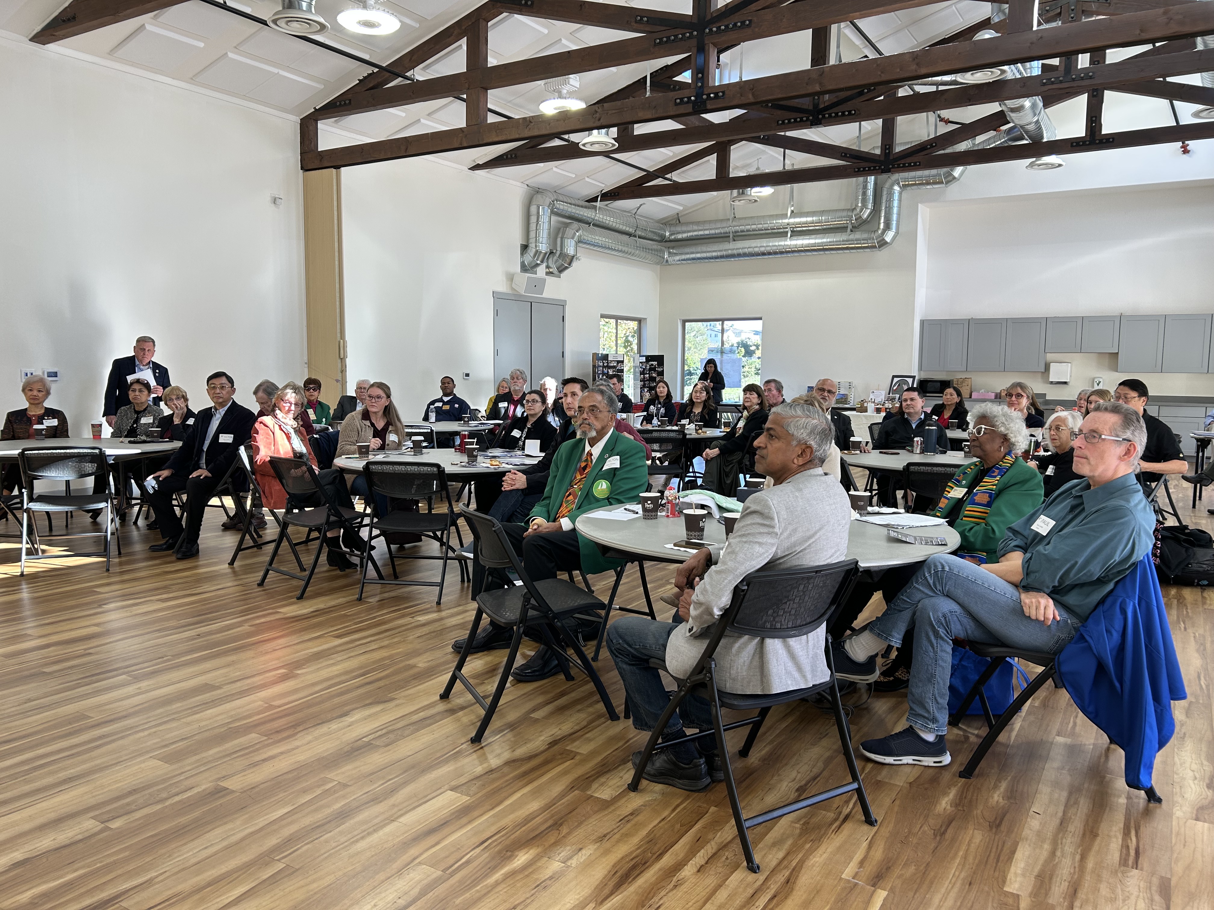 People sit at round tables in a bright room with exposed beams, attentively watching a presentation or speaker out of frame.