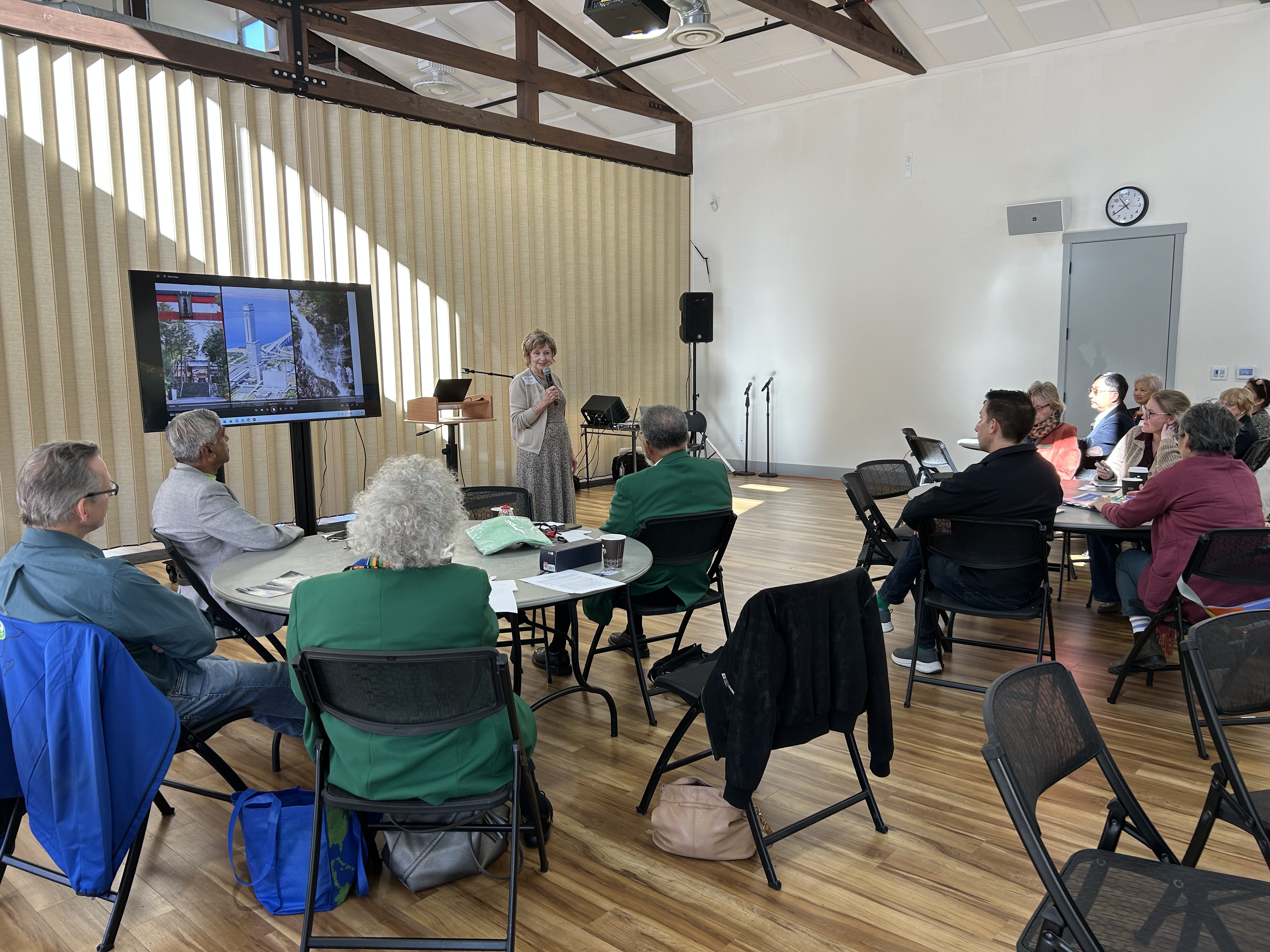 A group of people seated at round tables listen to a woman giving a presentation with slides displayed on a large screen in a well-lit room.