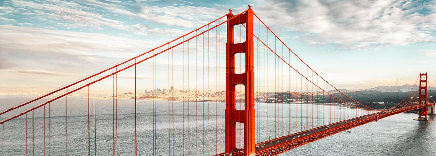 The Golden Gate Bridge spans across the water with the San Francisco skyline visible in the background under a partly cloudy sky.