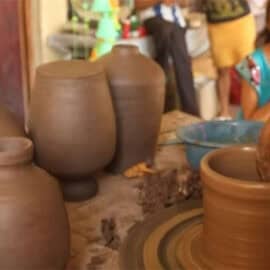 A person shapes clay on a pottery wheel, with several finished clay pots nearby—some destined for pottery sales—as a child watches in the background.