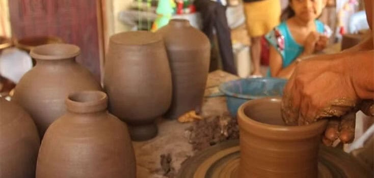 A person shapes clay on a pottery wheel, with several finished clay pots nearby—some destined for pottery sales—as a child watches in the background.
