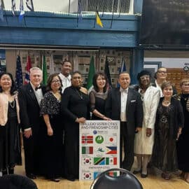 A group of people dressed in formal attire stand indoors, posing for a photo behind a sign listing sister and friendship cities with various international flags at the Vallejo Sister City Association Black & White Gala, celebrating community and culture.