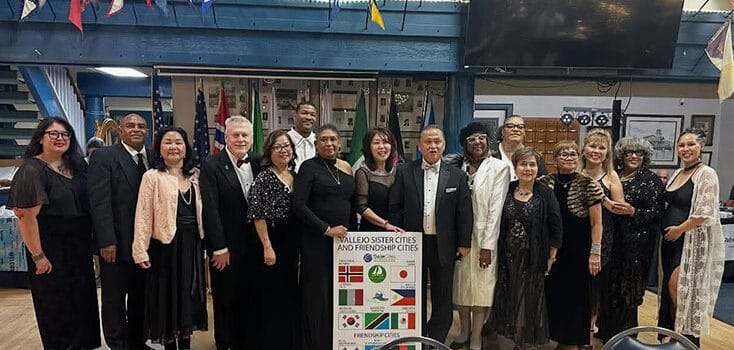 A group of people dressed in formal attire stand indoors, posing for a photo behind a sign listing sister and friendship cities with various international flags at the Vallejo Sister City Association Black & White Gala, celebrating community and culture.