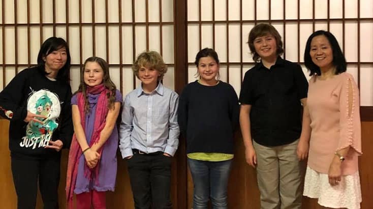 Six people, including four children and two adults, stand in a row and smile in front of a wooden panel wall with vertical and horizontal lines, participating in the Oakland-Fukuoka Youth Exchange youth exchange program.