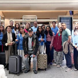 A group of Cupertino students with luggage pose for a photo in an airport terminal, ready for their exchange trip. A small white dog is near the front, and flight information displays are visible in the background.