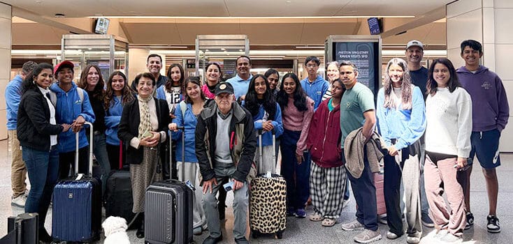 A group of Cupertino students with luggage pose for a photo in an airport terminal, ready for their exchange trip. A small white dog is near the front, and flight information displays are visible in the background.