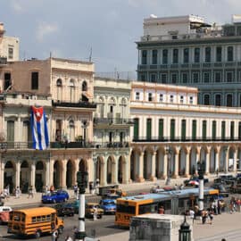 A busy street in Havana, Cuba with colorful colonial buildings, Cuban flags, cars, buses, and people walking on the sidewalks.