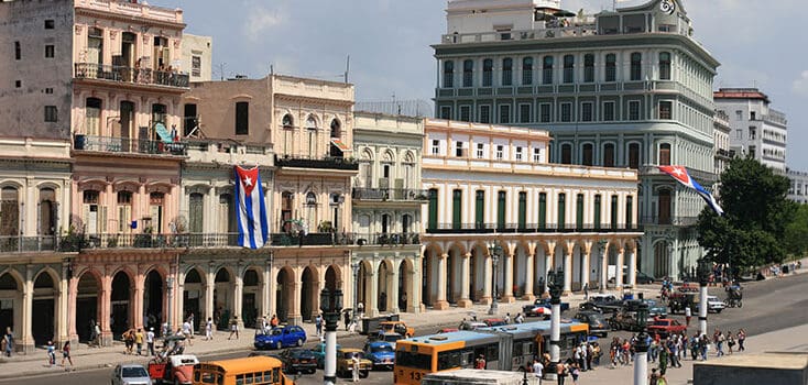 A busy street in Havana, Cuba with colorful colonial buildings, Cuban flags, cars, buses, and people walking on the sidewalks.