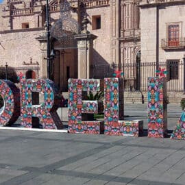 Large decorative letters spelling "MORELIA" with colorful patterns are displayed in a public square in front of historic stone buildings, highlighting the sister city partnership between Morelia Mexico and the Sacramento delegation.