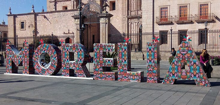 Large decorative letters spelling "MORELIA" with colorful patterns are displayed in a public square in front of historic stone buildings, highlighting the sister city partnership between Morelia Mexico and the Sacramento delegation.