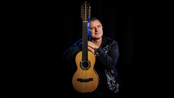 A man in a dark, embroidered jacket rests his arms on a 12-string acoustic guitar, reminiscent of traditional string instruments from Madeira, against a black background.