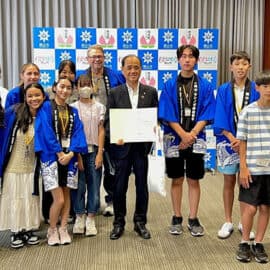 A group of young people and adults, including high school students from a Japan homestay in Okayama, pose indoors—some in blue robes holding certificates—in front of a backdrop with logos.
