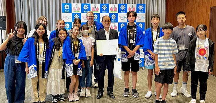 A group of young people and adults, including high school students from a Japan homestay in Okayama, pose indoors—some in blue robes holding certificates—in front of a backdrop with logos.