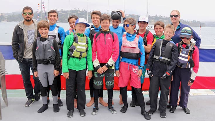 A group of teens and two adults in sailing gear pose for a photo on a dock in Sausalito, with water and boats behind them, capturing the spirit of Youth Sailing.