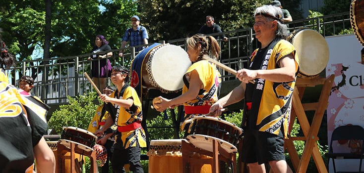 A group of people in traditional clothing play taiko drums outdoors during a cultural exchange event, with camellias blooming and a lively crowd among the trees in the background.