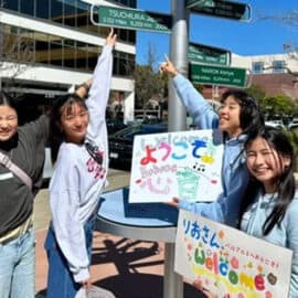 Four young women stand outdoors by a signpost—two are pointing upward, while two hold colorful welcome signs with Japanese writing, celebrating new friendships and connections through the Tsuchiura Exchange.