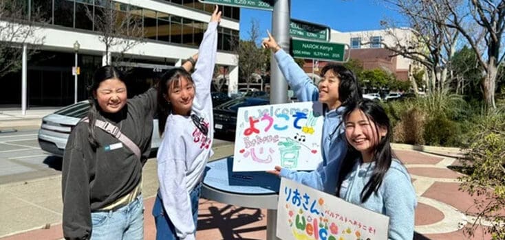 Four young women stand outdoors by a signpost—two are pointing upward, while two hold colorful welcome signs with Japanese writing, celebrating new friendships and connections through the Tsuchiura Exchange.