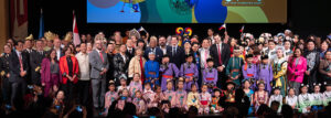 A large group of adults and children in formal and traditional attire pose for a group photo on stage at a cultural event.