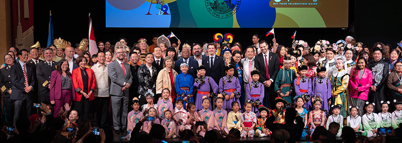 A large group of adults and children in formal and traditional attire pose for a group photo on stage at a cultural event.
